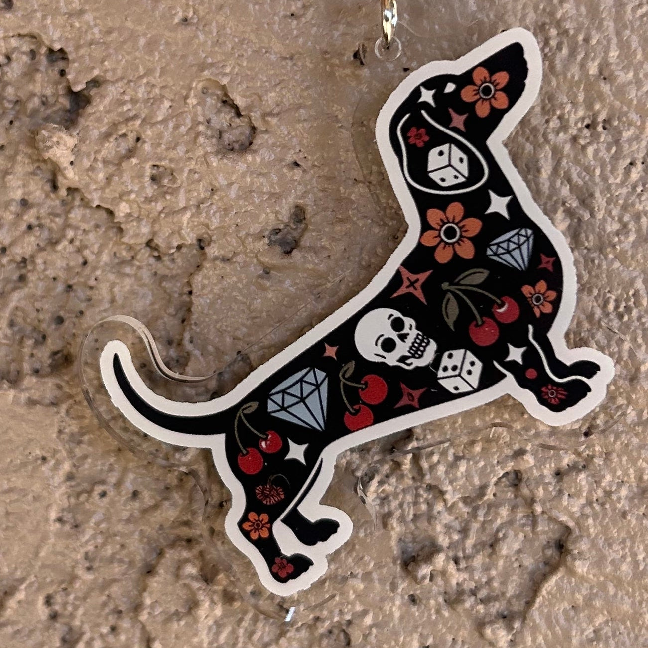 Keychain shaped like a dachshund with skull and crossbones design held by a hand on a textured surface.