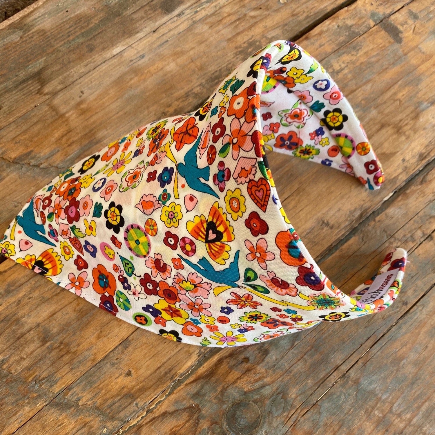 Head scarf that features brightly colored flowers and sparrow birds throughout the print. Has a white background. The scarf has been added to a headband for easy no-tie styling.