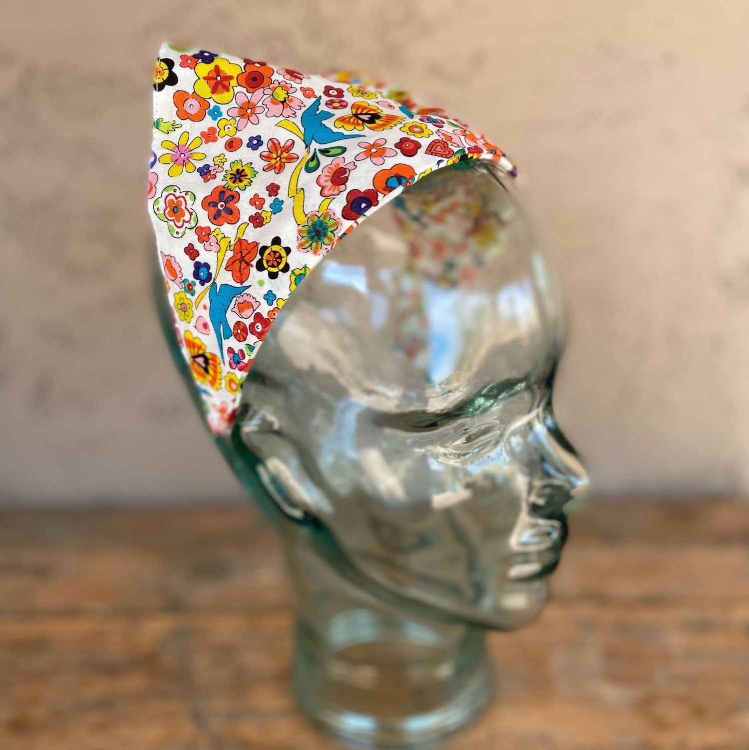 Head scarf that features brightly colored flowers and sparrow birds throughout the print. Has a white background. The scarf has been added to a headband for easy no-tie styling.