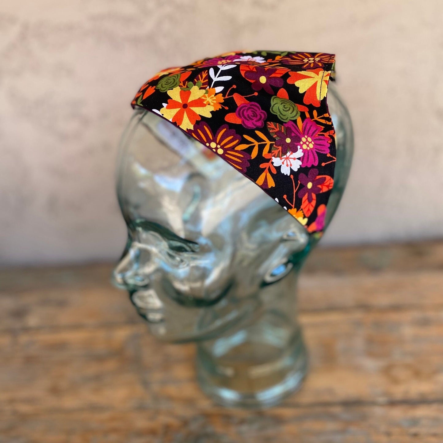 Triangle head scarf that has been attached to a headband. The print is a colorful floral design with orange, pinks , yellow and burgundy colors. Black background.